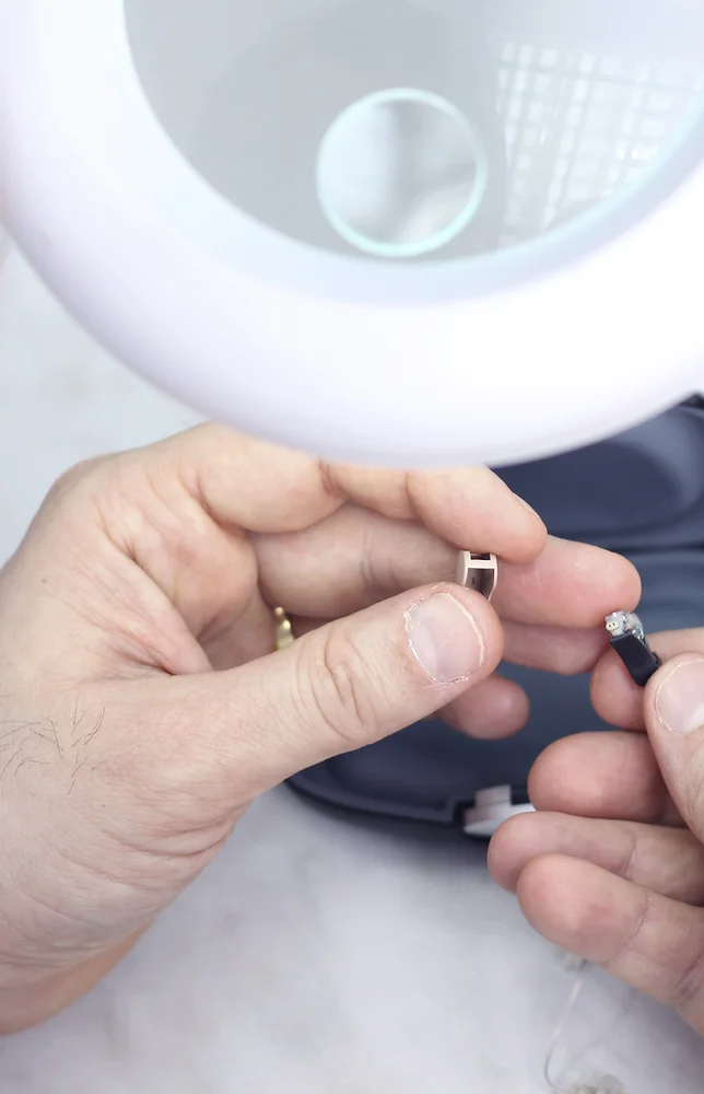 Two hands repair a hearing aid under a magnifying glass